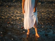 Portrait of crop charming little girl in white dress standing in water on beach — Stock Photo