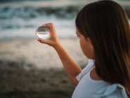 Little female child with brown hair holding glass ball on background of waves — Stock Photo