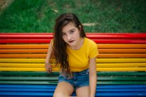 Jolie jeune femme en tenue décontractée souriant joyeusement et regardant la caméra tout en étant assis sur le banc arc-en-ciel dans le parc — Photo de stock