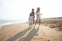 Niños felices y sonrientes en ropa casual corriendo descalzos a lo largo de la orilla del mar en la playa de arena en el día soleado de verano - foto de stock