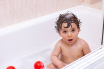 Bebê adorável olhando para a câmera com cabelo molhado enquanto toma um banho no banheiro brincando com brinquedos — Fotografia de Stock
