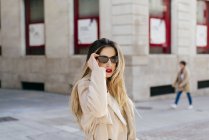 Young pretty female in stylish coat and sunglasses posing on street against marble building with bright red windows — Stock Photo