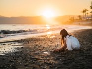 Cute pensive female kid in white dress playing with sand on seaside at sunset — Stock Photo