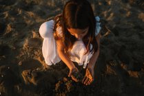 Bonito garoto feminino pensativo em vestido branco brincando com areia na praia à luz do sol — Fotografia de Stock