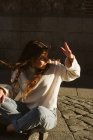Jeune femme en tenue décontractée souriant et couvrant le visage de la lumière du soleil assis sur le trottoir dans la rue de la ville et regardant loin — Photo de stock