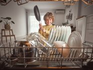 Annoyed little boy yelling while putting frying pan into open dishwasher in kitchen — Stock Photo