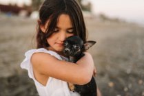 Portrait of charming female child in white dress holding little dog while sitting on sand — Stock Photo