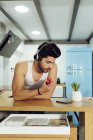 Jovem macho bonito com penteado elegante no fone de ouvido inclinado na mesa na cozinha e segurando maçã madura enquanto olha para o telefone — Fotografia de Stock