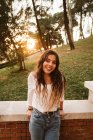 Jolie jeune femme en tenue décontractée souriant et regardant la caméra tout en se penchant sur la frontière par une journée ensoleillée dans le parc — Photo de stock
