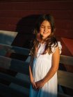 Ritratto di graziosa bambina in abito bianco appoggiata alla parete di legno alla luce del sole — Foto stock