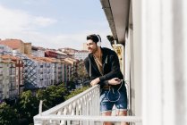 Junge lächelnde gutaussehende lässige männliche Person im Headset lehnt mit Smartphone an der Terrassenbrüstung und schaut weg — Stockfoto