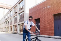 Молодой улыбающийся черный успешный мужчина с африканскими волосами в повседневной одежде и стильные солнечные очки, стоящие с велосипедом на улице — стоковое фото