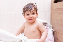 Liebenswertes Baby blickt mit nassen Haaren in die Kamera, während es nach der Dusche auf einem Handtuch im Badezimmer sitzt — Stockfoto