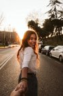 Menina bonita em roupa casual sorrindo e olhando para a câmera enquanto segurava a mão de pessoa irreconhecível na rua da cidade ao pôr do sol — Fotografia de Stock