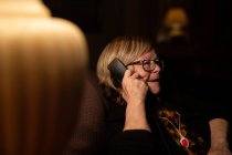 Mulher idosa feliz sorrindo e respondendo telefonema enquanto sentado no quarto escuro à noite em casa — Fotografia de Stock