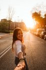 Menina bonita em roupa casual sorrindo e olhando para a câmera enquanto segurava a mão de pessoa irreconhecível na rua da cidade ao pôr do sol — Fotografia de Stock