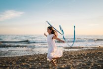 Маленькая девочка в белом платье бежит вдоль берега моря и держит длинную синюю ленту на фоне вечернего неба — стоковое фото