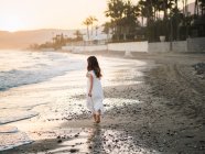 Vista laterale della bambina in abito bianco che cammina sulla riva del mare sullo sfondo del sole — Foto stock