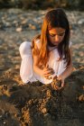 Cute pensive female kid in white dress playing with sand on beach in sunlight — Stock Photo