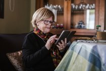 Старша жінка дивиться і зворушливий екран смартфона, сидячи на дивані у вітальні — стокове фото