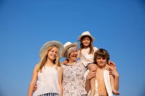 Abuela sonriente y nietos con sombreros y ropa de verano de pie juntos en un día soleado - foto de stock
