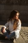 Jovem do sexo feminino em roupa casual sentado na calçada na rua da cidade e olhando para longe — Fotografia de Stock