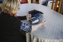 Seniorin misst mit Tensiometer Blutdruck, während sie zu Hause am Tisch sitzt — Stockfoto