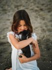 Retrato de encantadora criança feminina em vestido branco segurando pouco cão enquanto sentado na areia — Fotografia de Stock