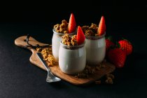 Glasses of cold tasty milk and delicious granola with strawberries on wooden board on black background — Stock Photo