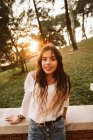 Hübsche junge Frau in lässigem Outfit lächelt und blickt in die Kamera, während sie sich an einem sonnigen Tag im Park an den Rand lehnt — Stockfoto