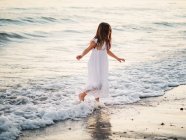 Little girl in white dress walking in water on beach — Stock Photo