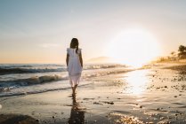 Niña en vestido blanco caminando en la orilla del mar en el fondo de la puesta del sol - foto de stock