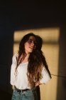 Молода жінка в повсякденному вбранні спирається на стіну будівлі в місці сонячного світла і дивиться в сторону — стокове фото