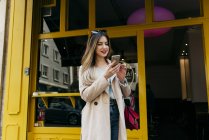 Joven mujer elegante de pie cerca de la cafetería con teléfono inteligente - foto de stock