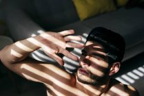 Зверху молодий голий красивий чоловік зі стильною зачіскою, що стоїть біля дивана і покриває сонячне світло рукою — стокове фото