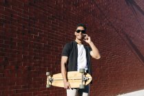 From below young African American cheerful handsome man in casual clothes and stylish sunglasses with long board speaking on phone — Stock Photo