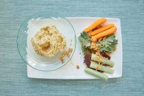 Зверху планування здорових натуральних овочів, таких як морква, огірок, салат і миска смачної гумусу на білій тарілці — стокове фото