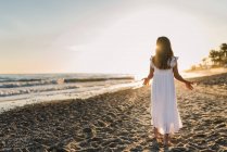 Kleines Mädchen in weißem Kleid, das am Ufer des Meeres im Hintergrund des Sonnenuntergangs spaziert — Stockfoto