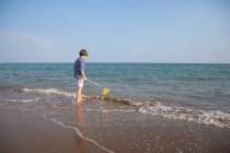 Vista lateral do cabelo longo bonito menino no verão desgaste de pé na água com rede de pesca na costa do mar em dia ensolarado — Fotografia de Stock