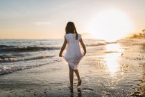 Niña en vestido blanco caminando en el agua en la playa en el fondo del sol - foto de stock