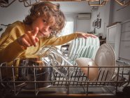 Мальчик улыбается и указывает на камеру, глядя в открытую посудомоечную машину на кухне — стоковое фото