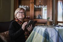 Старша жінка дивиться і зворушливий екран смартфона, сидячи на дивані у вітальні — стокове фото