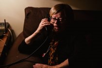 Heureuse femme âgée souriant et répondant à un appel téléphonique alors qu'elle était assise dans une pièce sombre le soir à la maison — Photo de stock