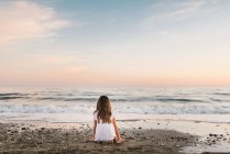 Back view of cute female child in white dress sitting on sandy seashore at sunset — Stock Photo