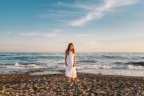 Portrait of charming little girl in white dress posing on sandy beach — Stock Photo