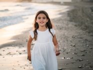 Menina bonita alegre no vestido branco andando na praia arenosa e olhando para a câmera — Fotografia de Stock