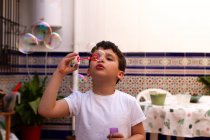 Маленький мальчик в белой футболке надувает мыльные пузыри, стоя дома на террасе — стоковое фото