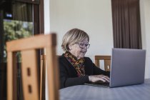 Mulher idosa em óculos navegando laptop enquanto sentado à mesa em casa aconchegante — Fotografia de Stock