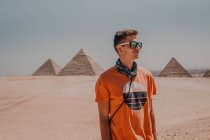 Fiducioso viaggiatore maschile in occhiali da sole guardando lontano mentre in piedi nel deserto contro le famose Grandi Piramidi al Cairo, Egitto — Foto stock