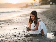 Mignon enfant féminin coûteux en robe blanche jouant avec le sable sur le bord de la mer au soleil — Photo de stock